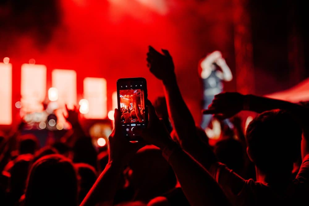 A fan holding up a phone to film the band through red stage light and haze